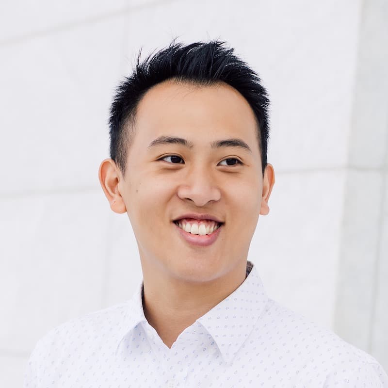 Portrait of a person with short black hair wearing a white button-down shirt smiling in front of a white wall.